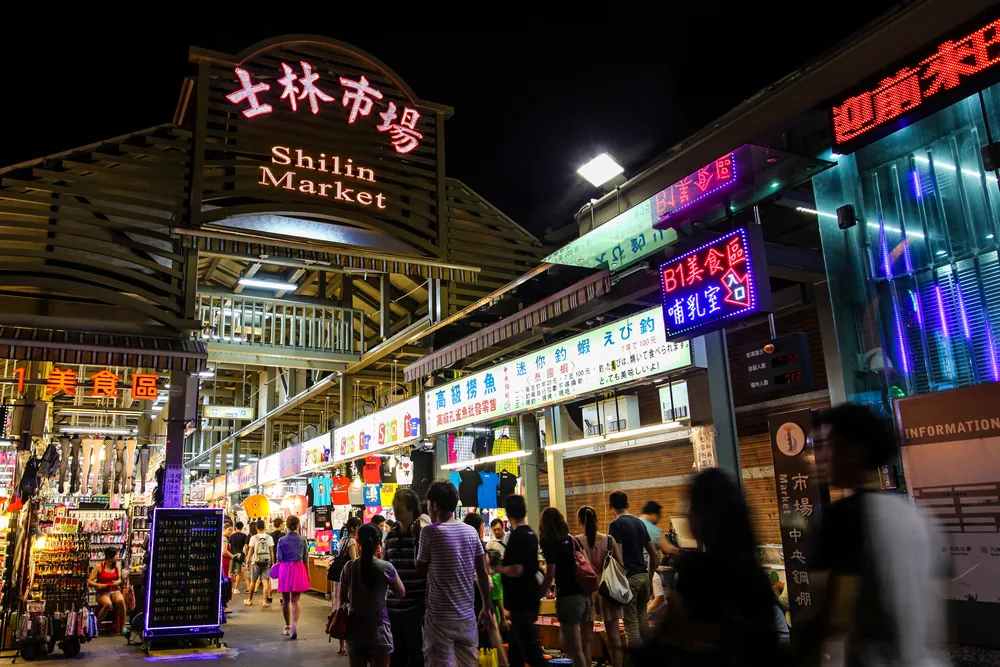 Shilin Night Market bustling with crowds beneath neon signs, vendor stalls offering street food and merchandise in all directions.